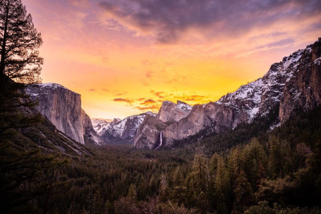 Quintessential view of Bridal Veil Falls, El Capitan, and Half Dome in the back from Tunnel View on a beautiful sunrise morning with orange and pink skies illuminating the beginning of the day.